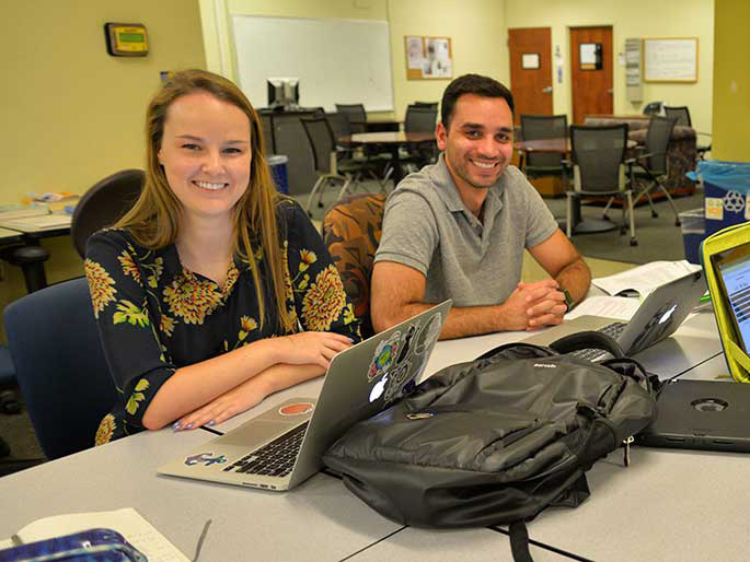 Two students sit at a table with open laptops